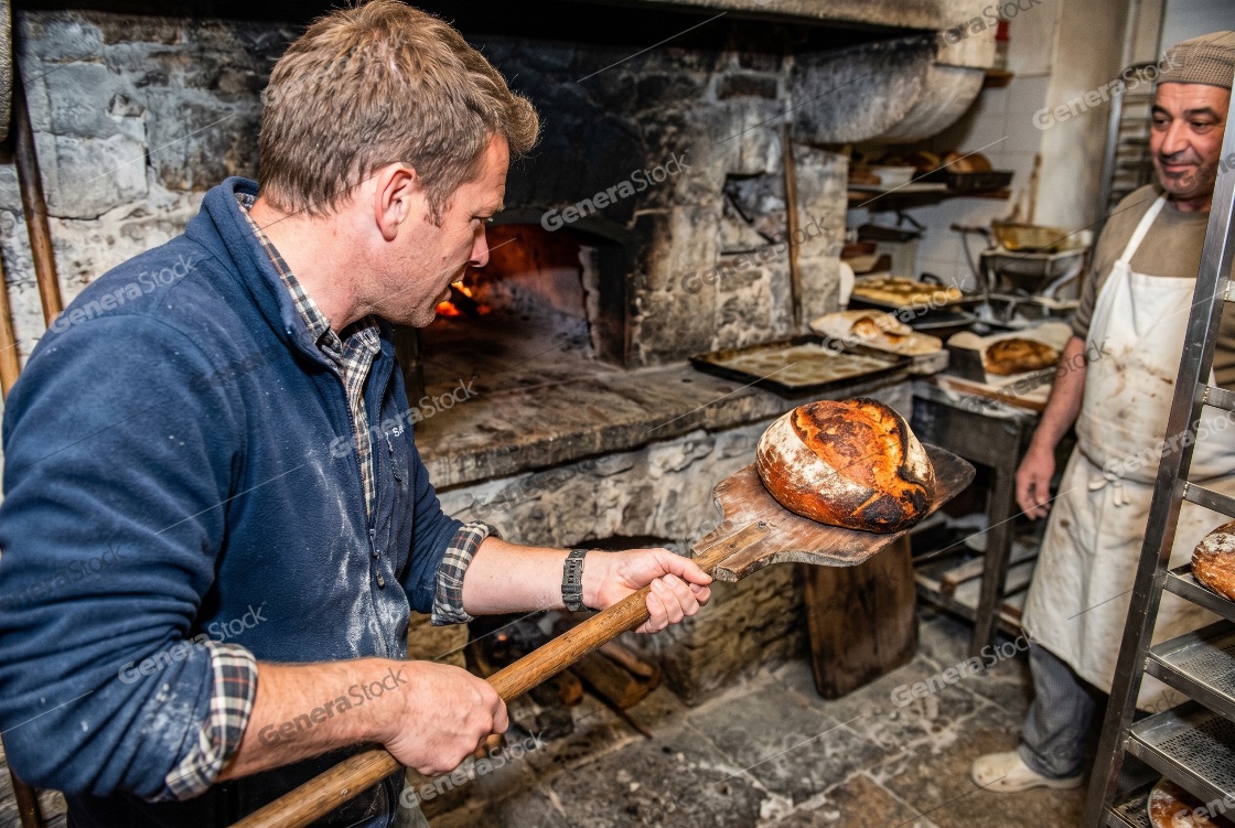 Male baker removing a fresh loaf of artisan sourdough bread from a traditional wood-fired stone oven in a rustic bakery