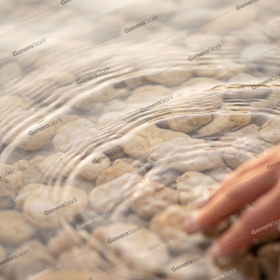 A close-up shot of a person's hand gently touching the surface of clear, shallow water, creating soft ripples over smooth white pebbles at the bottom during a warm, golden sunset