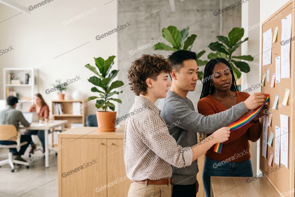Diverse team of young professionals pinning a rainbow ribbon to a corkboard in a bright modern office, demonstrating workplace inclusion and pride support