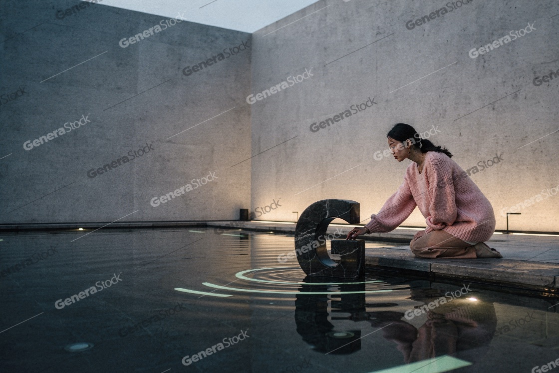 Young Asian Woman Kneeling Beside Reflecting Pool Touching Marbl