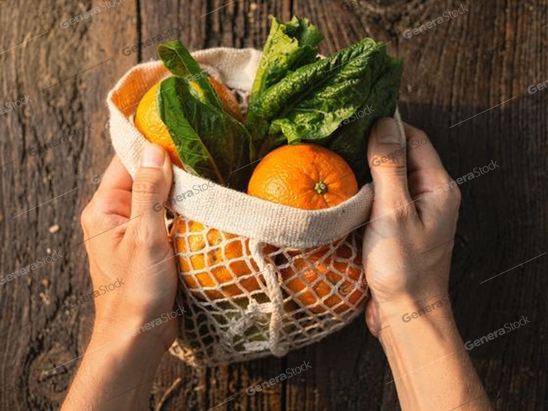 hands holding a cotton mesh bag with oranges and chard, dark wood