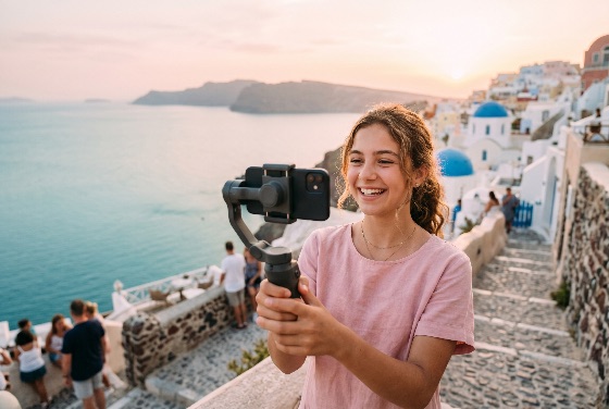 happy, young, woman, recording, travel, vlog, with, smartphone, and, gimbal, stabilizer, oia, santorini, greece, during, beautiful, sunset, vlogger, vacation, influencer, content, creator, smiling, technology, social, media, aegean, sea, tourism, island, mediterranean, cyclades, white, houses, blue, dome, scenic, landscape, golden, hour, summer, holiday, adventure, lifestyle, filming, people