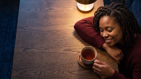high, angle, view, relaxed, smiling, african, american, woman, with, top-down, shot, black, dreadlocks, resting, her, head, folded, arm, dark, wooden, table, she, wears, cozy, burgundy, sweater, and, holds, hot, cup, tea, saucer, soft, warm, light, from, lamp, creates, serene, atmosphere, ample, copy, space, the, left, mug, relaxing, happy, drink, beverage, rest, peaceful, indoor, lifestyle, comfort, tranquility, break, coffee, calm, satisfaction, contentment, adult, female, leisure, top, home, hairstyle, wood, surface, grain, cheerful, time, night, glowing, people