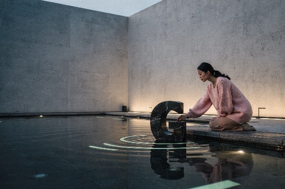 young, asian, woman, kneeling, beside, reflecting, pool, touching, marbl, east, soft, pink, sweater, kneels, the, edge, sleek, modern, she, gently, touches, large, black, marble, letter, sculpture, partially, submerged, water, causing, delicate, ripples, across, surface, scene, set, elegant, minimalist, architectural, space, with, smooth, concrete, walls, and, warm, ground, lighting, creating, calm, reflective, contemporary, atmosphere, architecture, ripple, serene, peaceful, luxury, fashion, reflection, tranquility, courtyard, design, art, aesthetic, stone, stylish, lifestyle, outdoor, building, zen, artistic, concept, quiet, wall, model, relaxation, harmony, structure, female, buildings