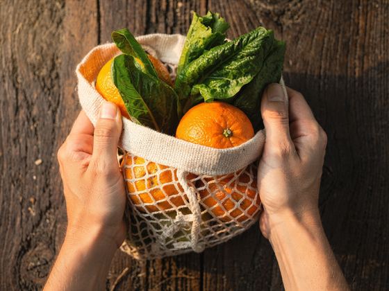 hands holding a cotton mesh bag with oranges and chard, dark wood